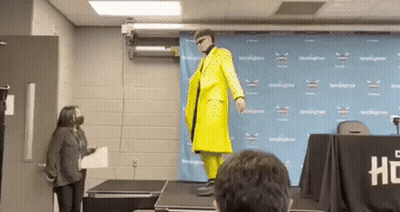 A Man In A Yellow Suit Is Standing On A Stage In Front Of A Crowd . GIF