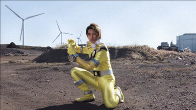 A Man In A Yellow Superhero Costume Is Kneeling In A Dirt Field GIF