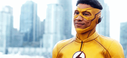 A Man In A Yellow Superhero Costume With A Lightning Bolt On His Chest Stands In Front Of A City Skyline . GIF