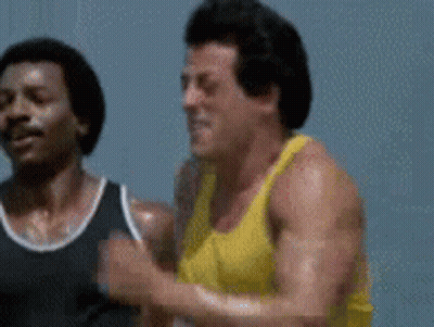 A Man In A Yellow Tank Top Is Running Next To Another Man . GIF