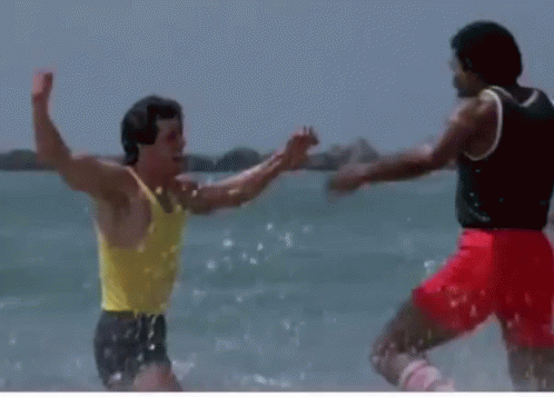 A Man In A Yellow Tank Top Is Standing Next To Another Man In Red Shorts On The Beach GIF