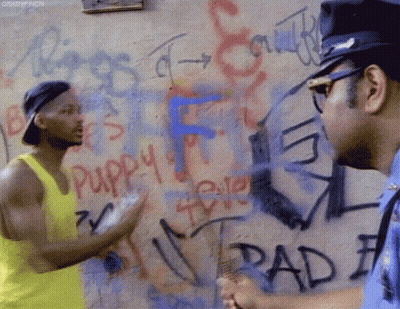 A Man In A Yellow Tank Top Talks To A Police Officer In Front Of A Wall Covered In Graffiti Including The Word Puppy GIF