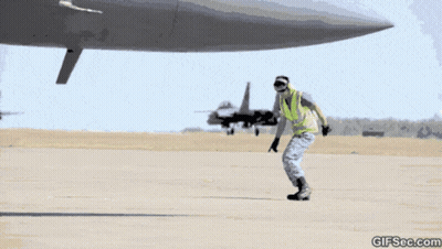 A Man In A Yellow Vest Is Standing In Front Of An Airplane With A Gifsec.com Watermark GIF