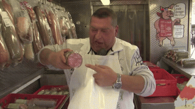 A Man In An Apron Holds A Piece Of Meat In Front Of A Sign That Says " Die Größte Europäische Wurst " GIF