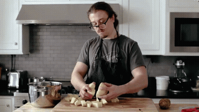 A Man In An Apron Is Peeling Potatoes On A Wooden Cutting Board GIF
