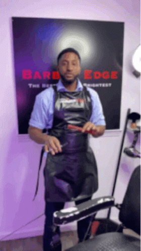 A Man In An Apron Is Standing In Front Of A Barber Chair Holding A Comb . GIF