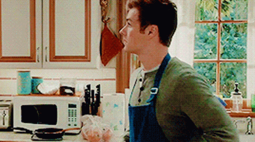 A Man In An Apron Stands In A Kitchen GIF