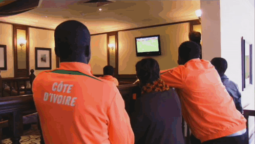 A Man In An Orange Cote D' Ivoire Jacket Watches A Game GIF