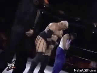 A Man In An Orange Hard Hat Is Standing Next To A Wrestler In A Ring . GIF