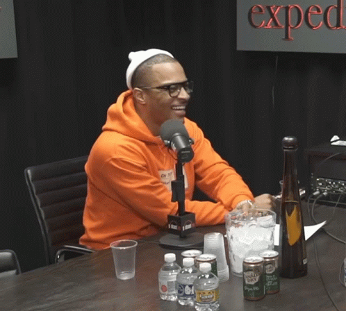 A Man In An Orange Hoodie Sits In Front Of A Microphone In Front Of A Sign That Says " Expect " GIF