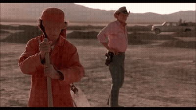 A Man In An Orange Jacket Is Holding A Shovel In The Desert While Another Man In A Pink Shirt Stands Behind Him . GIF