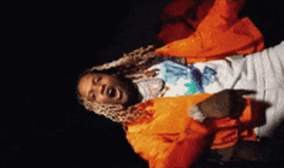 A Man In An Orange Jacket Is Singing Into A Microphone In A Dark Room . GIF