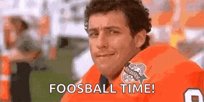 A Man In An Orange Jersey Is Sitting On A Football Field With The Words `` Fooball Time '' Written On His Face . GIF