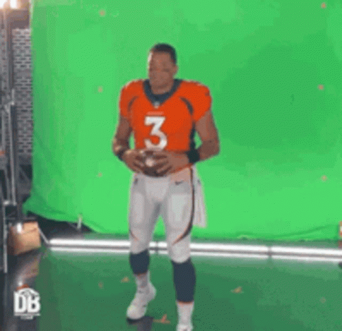 A Man In An Orange Jersey With The Number 3 On It Holds A Football In Front Of A Green Screen . GIF