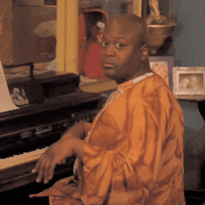 A Man In An Orange Robe Sits At A Piano With A Sign That Says S On It GIF