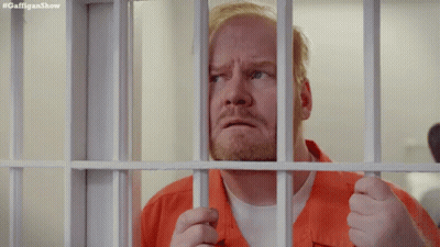 A Man In An Orange Shirt Is Behind Bars In A Prison Cell . GIF