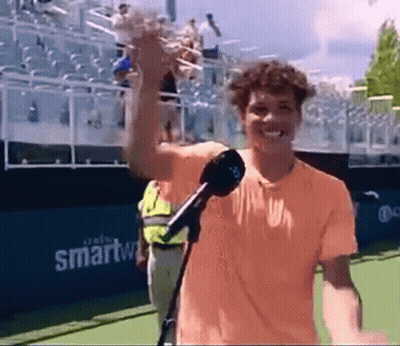 A Man In An Orange Shirt Is Holding A Microphone On A Tennis Court GIF
