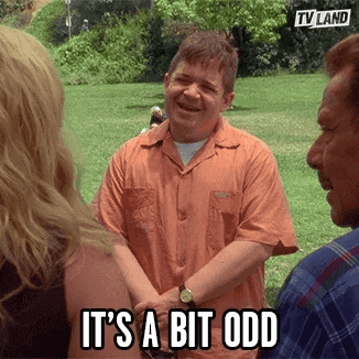 A Man In An Orange Shirt Is Standing In A Park Talking To A Group Of People And Laughing . GIF