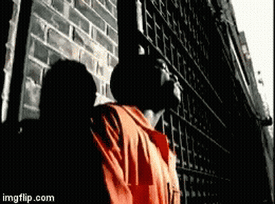 A Man In An Orange Shirt Is Standing In Front Of A Brick Wall . GIF