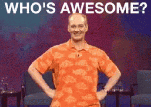 A Man In An Orange Shirt Is Standing On A Stage With His Hands On His Hips And The Words " Who 'S Awesome " Above Him GIF