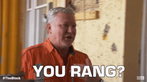 A Man In An Orange Shirt Says " You Rang " In Front Of A Yellow Wall GIF