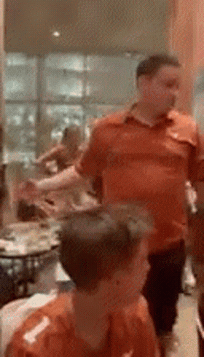 A Man In An Orange Shirt Stands In A Room With Other People GIF