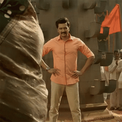 A Man In An Orange Shirt Stands With His Hands On His Hips In Front Of A Crowd GIF