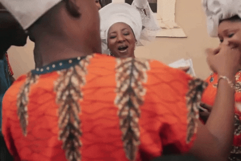 A Man In An Orange Shirt With Feathers On The Sleeves Is Standing Next To A Woman In A White Turban GIF
