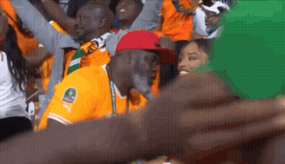 A Man In An Orange Shirt With The Letter C On It Is Sitting In A Crowd Of People . GIF