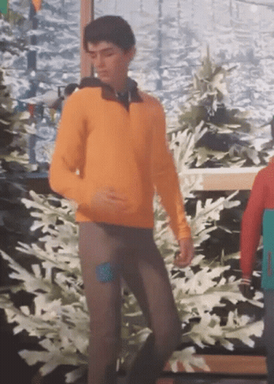 A Man In An Orange Sweater And Brown Pants Is Dancing In Front Of A Painting Of Snow Covered Trees . GIF