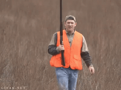 A Man In An Orange Vest Is Holding A Shotgun In A Field GIF