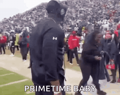 A Man In An Under Armour Jacket Is Standing On A Football Field With A Crowd Behind Him And Says Primetime Baby . GIF