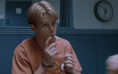 A Man In Orange Scrubs Is Sitting In Front Of A Wall Clock That Says Sounds Like GIF
