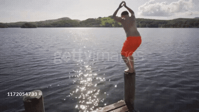 A Man In Orange Shorts Is Jumping Into A Lake From A Pier GIF