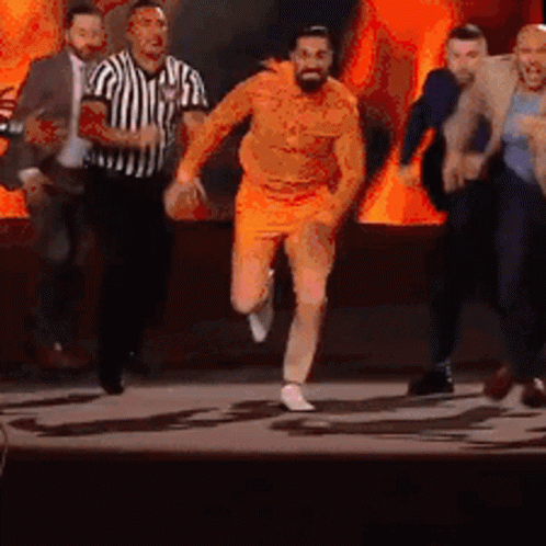 A Man In Orange Shorts Is Running On A Stage With A Referee Behind Him . GIF