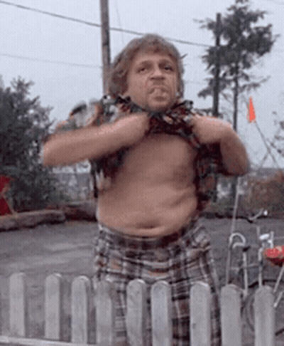 A Man In Plaid Shorts Is Standing In Front Of A Fence And Taking Off His Shirt GIF