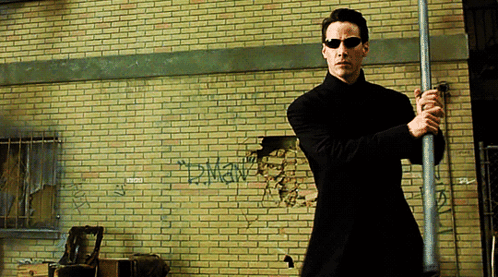 A Man In Sunglasses Holds A Pole In Front Of A Brick Wall With Graffiti On It That Says Dman GIF