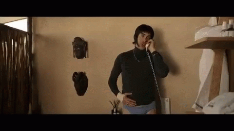 A Man In Underwear Is Talking On A Phone While Standing In A Room . GIF