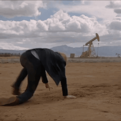 A Man Is Bending Over In The Dirt In Front Of A Oil Pump GIF