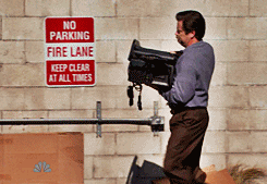 A Man Is Carrying A Camera In Front Of A Sign That Says No Parking Fire Lane Keep Clear At All Times GIF