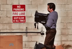 A Man Is Carrying A Suitcase In Front Of A No Parking Fire Lane Sign GIF