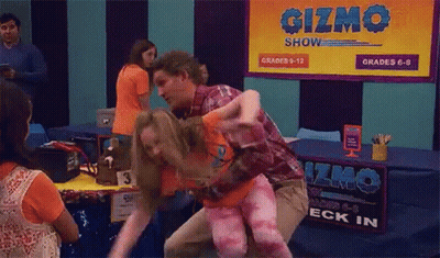 A Man Is Carrying A Woman In His Arms In Front Of A Sign That Says Gizmo Show . GIF