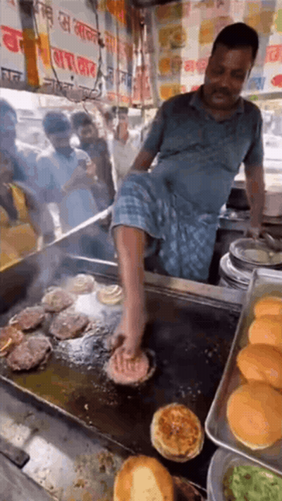 A Man Is Cooking Burgers On A Grill With His Foot GIF