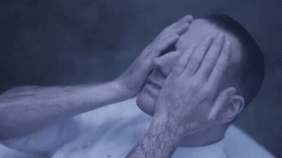 A Man Is Covering His Eyes With His Hands While Laying On A Bed . GIF