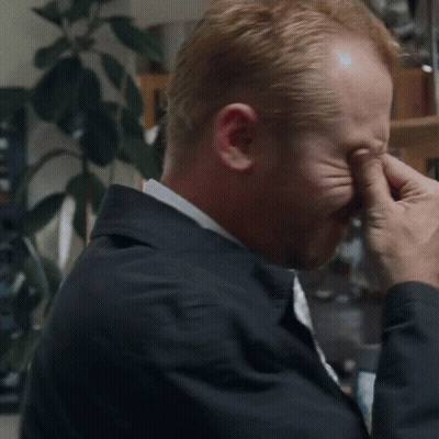 A Man Is Covering His Eyes With His Hands While Wearing A Jacket And Tie . GIF