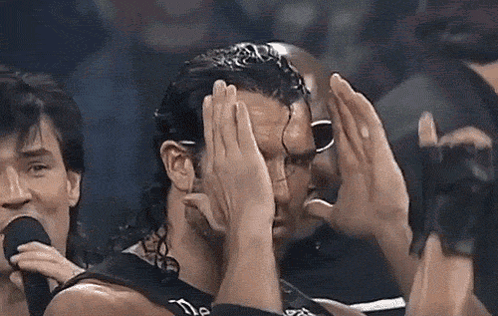A Man Is Covering His Face With His Hands While Another Man Holds A Microphone Behind Him . GIF