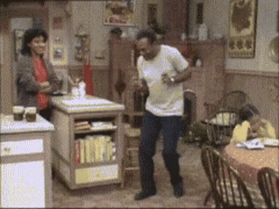 A Man Is Dancing In A Kitchen While A Woman Sits At A Table GIF