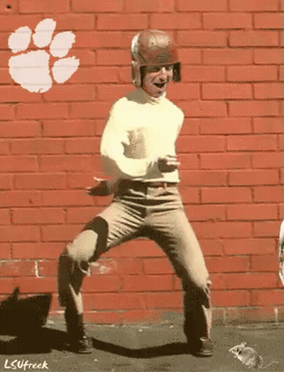 A Man Is Dancing In Front Of A Brick Wall That Has A Clemson Logo On It GIF