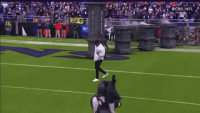 A Man Is Doing A Trick On A Football Field In Front Of A Ravens Sign GIF