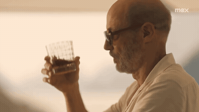 A Man Is Drinking A Glass Of Whiskey With A Max Logo Behind Him GIF